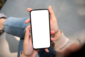 A close-up image of a woman in jeans sits outdoors on a sunny day and uses her smartphone.