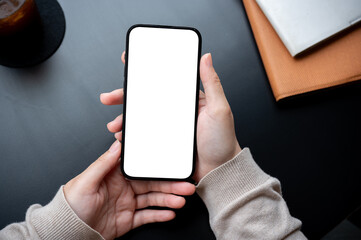 Top view image of a woman holding a smartphone white-screen mockup over an office desk workspace.