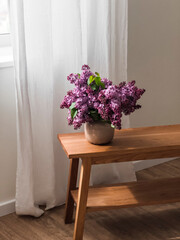 Fototapeta premium A bouquet of lilacs in a ceramic vase on a wooden oak bench in the living room