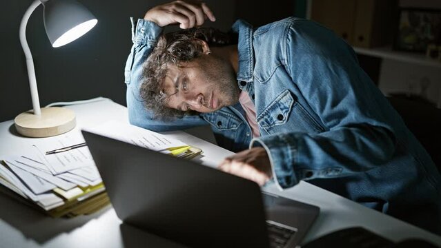 An overworked man sitting tiredly at his desk indoors, with a laptop, paperwork, and desk lamp in a dark office.
