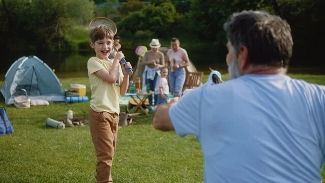 Slow motion shot of little boy playing bagminton with grandfather on nature camping with family for weekend or vacation on a meadow by river