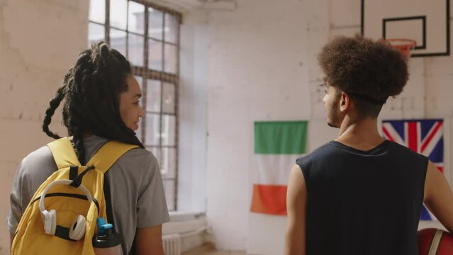 Tracking back-view slowmo of young Biracial couple of basketball players talking while walking along indoor court, arriving on training