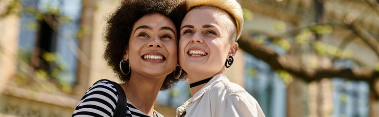 Two young women, a multicultural lesbian couple, share joyful smiles in front of a university...