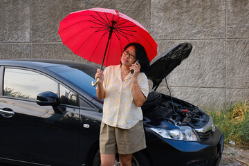 A woman under a red umbrella was checking the engine of her broken down car parked on the side of...