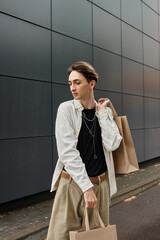 Fototapeta premium A young LGBTQ individual in stylish attire carrying shopping bags in front of a modern building.