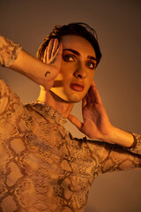 A young queer in a snake skin dress poses with hands on her ears in a stylish, pride-inspired...