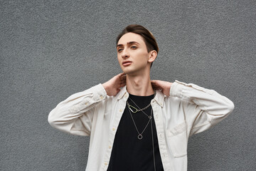 A young queer person confidently poses against a gray wall, showcasing his stylish attire with...