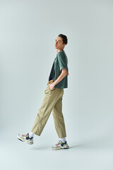 A young queer person confidently poses in a studio, wearing a stylish t-shirt and khaki pants on a grey background.