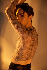 A young queer person in a striking snakeskin top poses in front of a mirror, exuding confidence and...