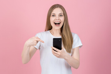 Attractive caucasian young blonde woman in casual white t-shirt, smiling, looking at camera, using and pointing finger at mobile phone isolated on pink studio background.