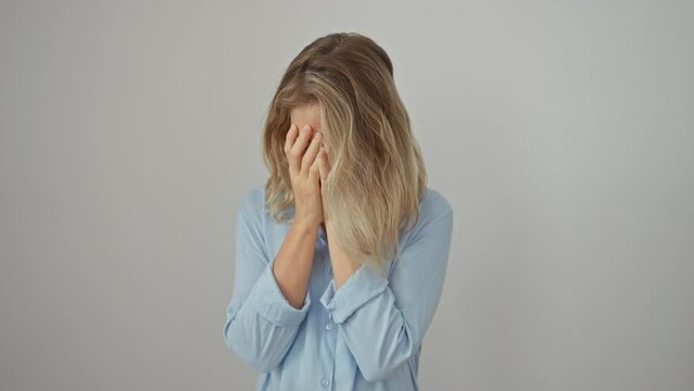 Despondent blonde young woman wearing shirt, stand alone against white isolated background, covering face with hands, crying out her worries and depression.
