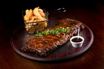 succulent pork ribs in barbecue sauce with basket of rustic potatoes and bbq sauce on wooden board...