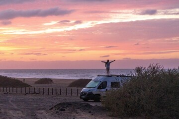 Embracing freedom and adventure: individual standing with arms outstretched on a van, silhouetted against a serene sunset by a tranquil coastal landscape