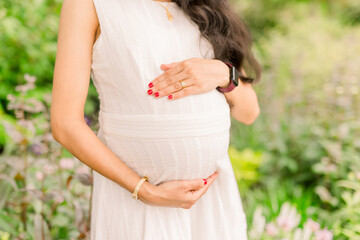 A pregnant woman in her third trimester wears a white dress outside in a garden and cradles her...