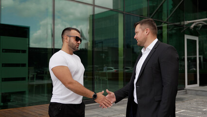 Businessmen have concluded an agreement against the backdrop of skyscrapers. A man in a suit and a man in a white T-shirt. Businessman and athlete shake hands.
