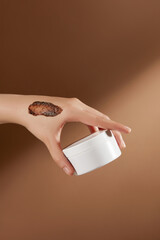 Frontal shot photo on brown background, a blank label cosmetic pot of exfoliation be held by a hand...