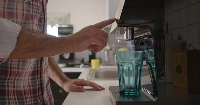 A man stands at a kitchen counter, pouring crystal clear water into a blue glass from a modern home water filtration system, with natural light illuminating the scene.