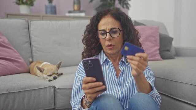 A middle-aged woman looks worriedly at her phone while holding a credit card, unaware of the sleeping chihuahua on the sofa in a cozy living room.
