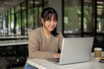 A young Asian female college student working remotely at a canteen, working on her laptop computer.