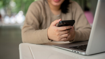 A young Asian woman using her smartphone at a table indoors, a laptop computer on the table.