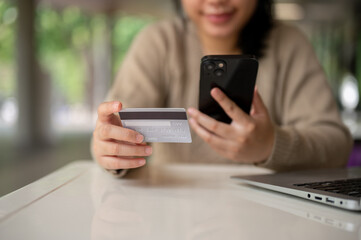 A young happy Asian woman using her mobile banking app while sitting at a table indoors.