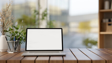 A front view of a laptop computer mockup and decorative objects are on a wooden tabletop in a room.
