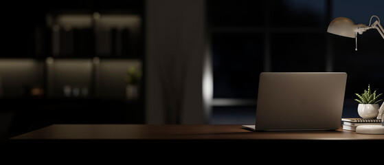 A laptop computer on a table in a modern dark room at night with a dim light from a table lamp.