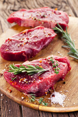 Raw beef steaks with herbs and spices ready for cooking are on wooden board.