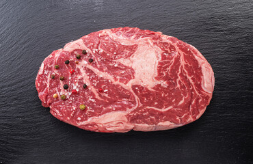 Raw ribeye steak with pepper corns and rosemary on graphite serving board, top view.