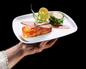 Chef holds white plate of  roasted salmon with side dish of green beans on black background. Tasty food background.