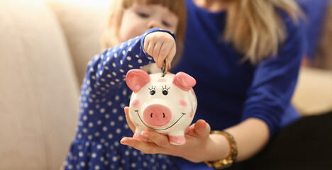 Child little girl arm putting pin money coins into happy pink faced piglet slot closeup. Making...