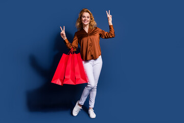Fototapeta premium Full length photo of adorable woman dressed silk shirt hold shopping bags showing v-sign symbol isolated on dark blue color background