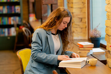 in bookstore near the window a beautiful student is sitting reading books preparing for exams