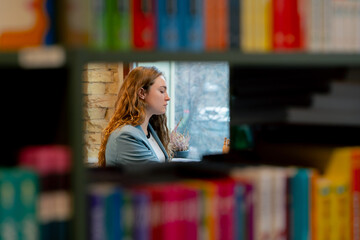 in bookstore near the window a beautiful student is sitting reading books preparing for exams
