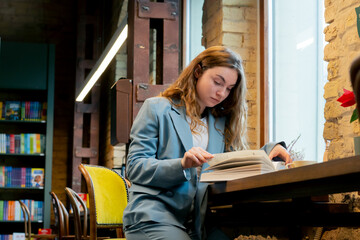in bookstore near the window a beautiful student is sitting reading books preparing for exams