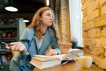 in bookstore near window a beautiful student is sitting boring reading books preparing for exams