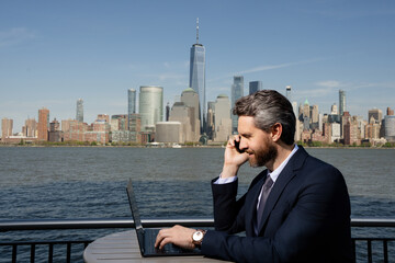 Business man using laptop and talking on the phone outdoor in NYC. American business success. Remote freelance online work. Online business. Outside business. Mature serious freelancer using phone.