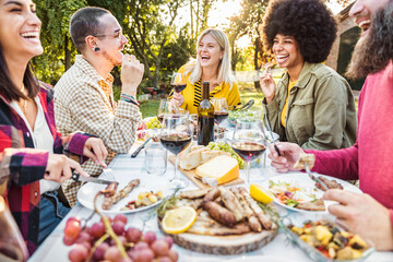 Happy friends having bbq dinner party in garden restaurant - Multiracial young people eating grill meat and drinking red wine in backyard - Food life style concept with guys and girls sitting outdoors