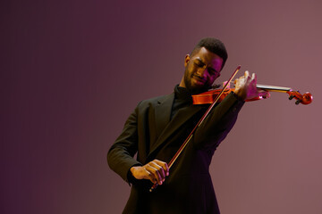 Elegant musician in black suit playing violin on vibrant purple background in studio setting