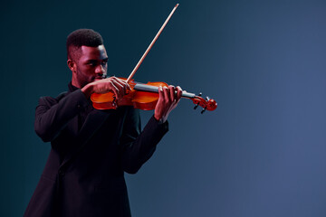 Soulful young African American man performing with violin against dark blue background in studio...
