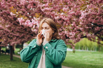 Sneezing young redhead woman with nose wiper among blooming trees in park. Portrait of sick women sneezes in white tissue, suffers from rhinitis and running nose. Symptoms of cold or allergy.