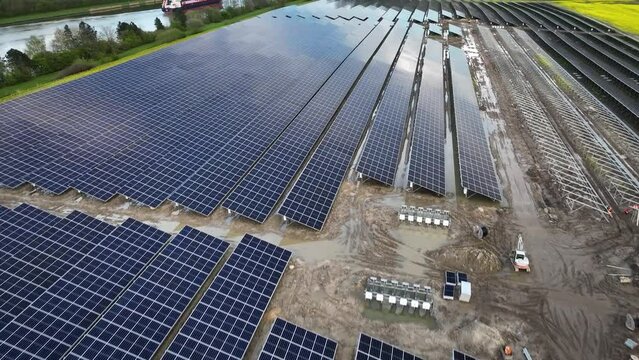 Shot of industrial solar power station, currently under construction, solar modules, array of white combiner boxes with power cables for connection situated near canal, where ship navigate waterway.  