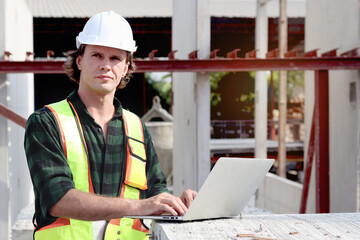 Portrait of happy worker engineer with safety vest and helmet typing on laptop computer while inspecting working area at construction building site. Young architect works at building site workplace.