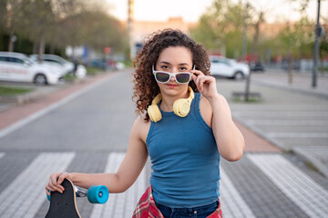 Fashionable young woman with curly hair, wearing sunglasses and headphones, holding a skateboard in an urban park, dressed in a blue tank top and plaid shirt. 