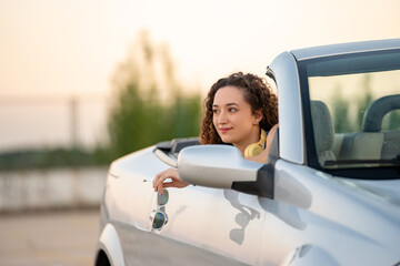 Curly-haired young woman enjoys a serene view from a convertible car during a tranquil sunset.