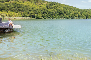 Young woman in white clothes is sitting on wooden pier and feeding wild ducks.