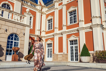 Fascinating young woman in long dress with floral pattern and staw hat is walking near hotel or...