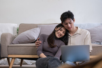 Fototapeta premium Young Asian couple in love Sitting happily together looking at laptop and relaxing in the living room. Couple making romantic love in the living room