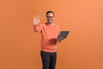 Cheerful handsome freelancer with laptop computer showing OK sign confidently on orange background
