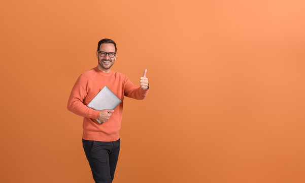 Portrait of happy male manager holding computer and showing thumbs up sign over orange background